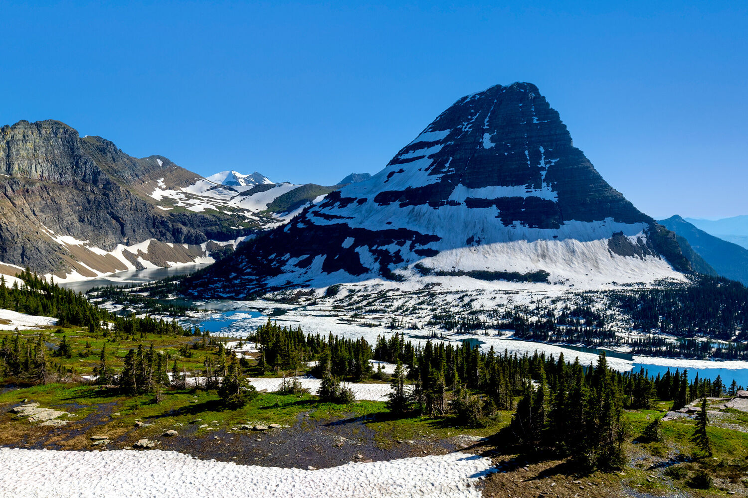 Hidden Lake begins to thaw in early July below the looming shadow of Bearhat Mountain.  In the distance, snow-covered Gunsight...