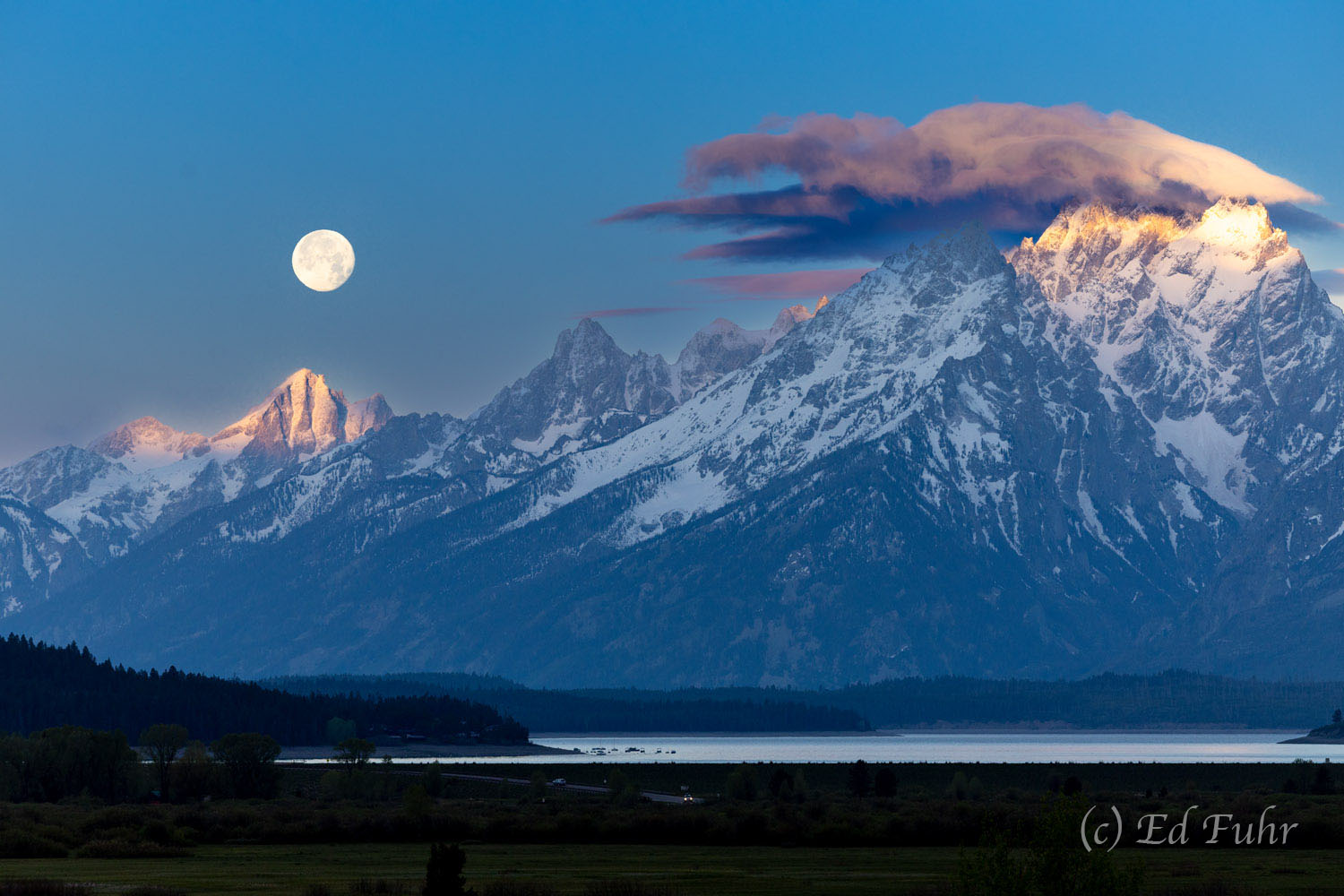 Sunrise has struck the high peaks of the Teton mountain range as a full moon begins to slip into a new day. Dwindling Lake Jackson...
