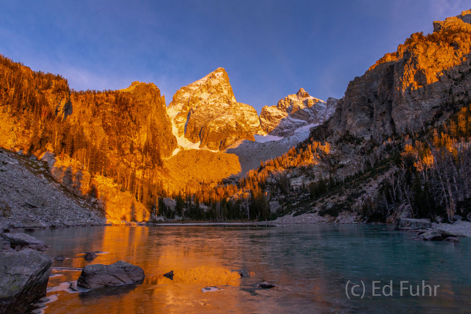 Coated in ice, a high alpine lake reflects the glow or morning's first light, below the peak of the Grand.