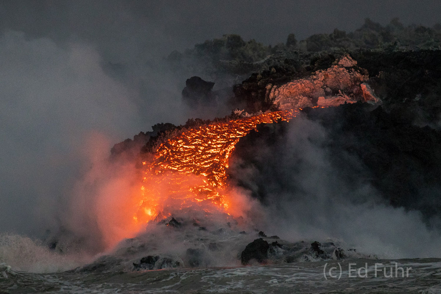 On one of our close sweeps by the shore, we see the torrent of molten lava flowing into the ocean where it cools and instantaneously...