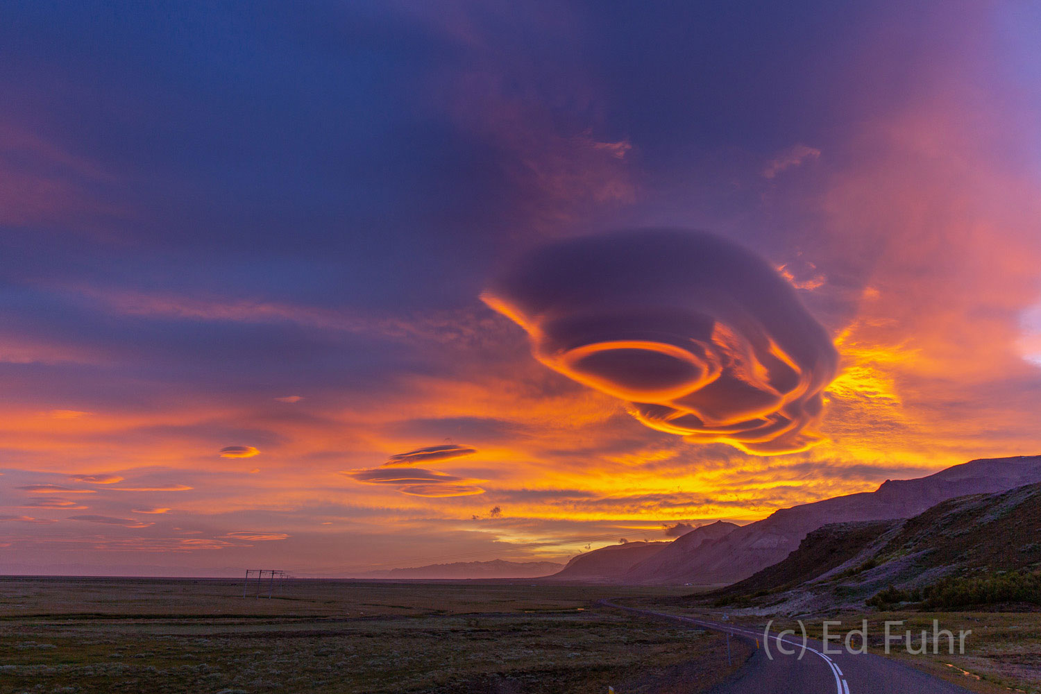 Streams of lenticular clouds float across the midnight sky, casting an eerie glow across the volcanic landscape.