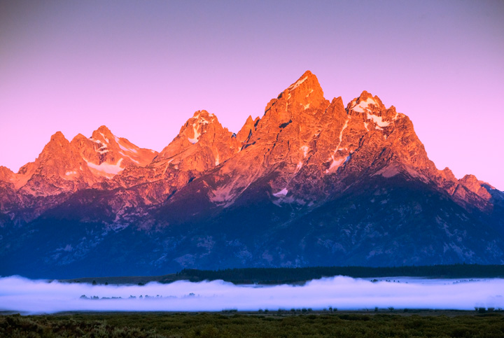 The sun bathes the Teton range above a bank of fog rising above the meandering Snake River that wends its way below.&nbsp;